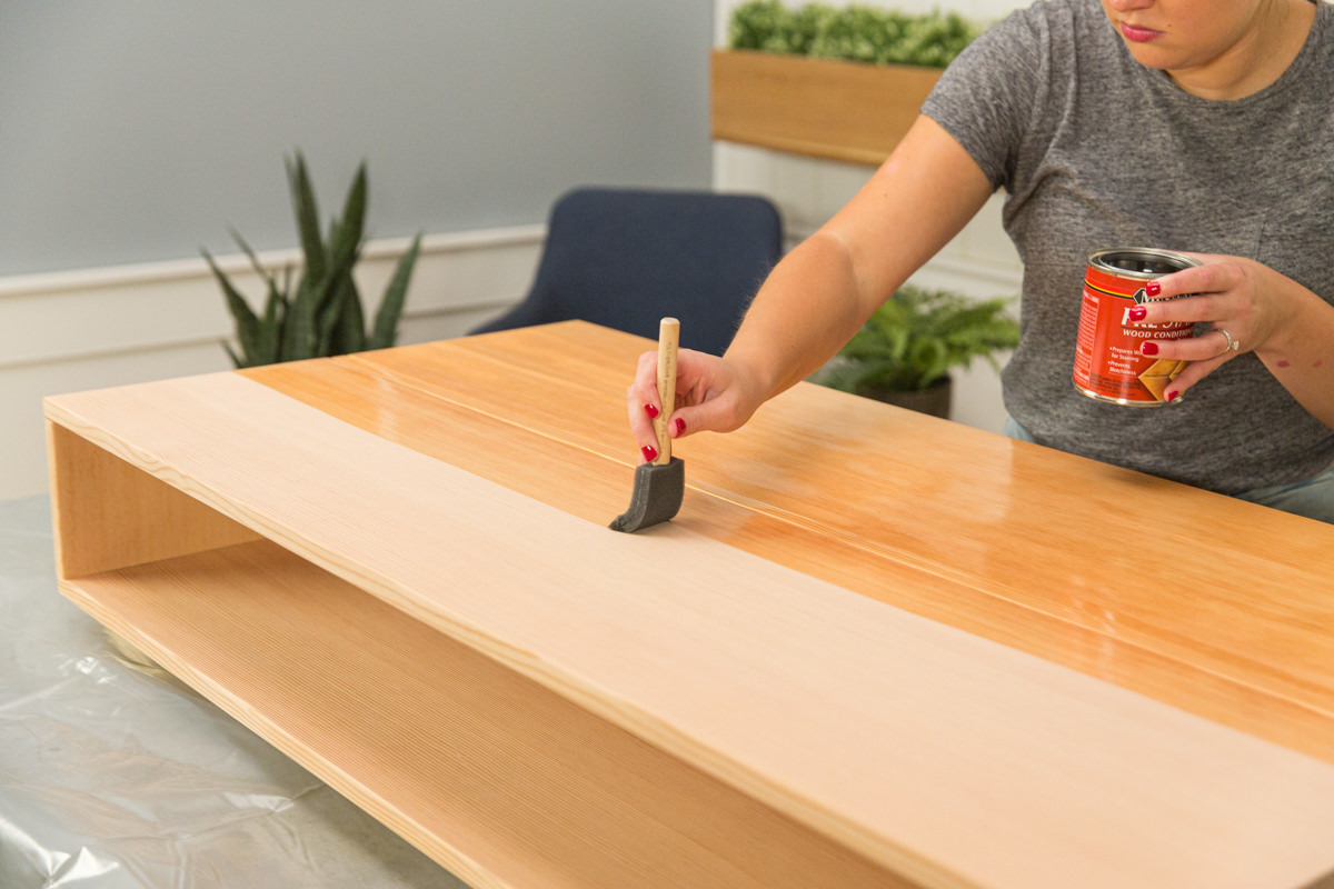 Applying wood conditioner to wood DIY coffee table assembly with foam brush.