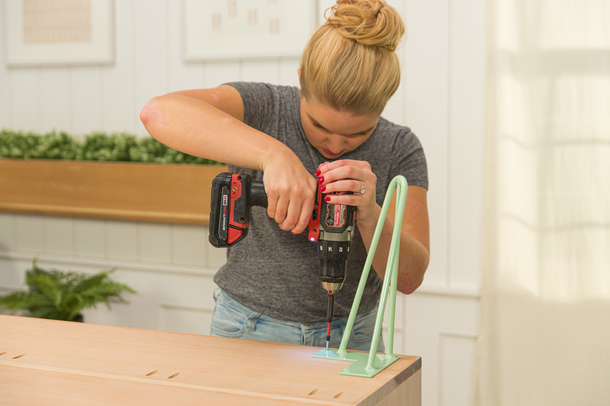 Driving screw with power drill to attach mint green hairpin leg to wood DIY coffee table assembly.