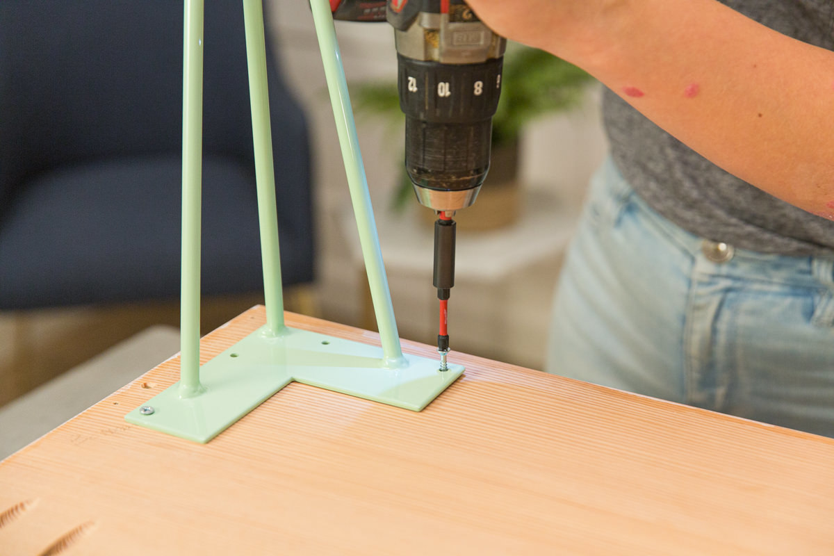 Driving screw with power drill to attach mint green hairpin leg to wood DIY coffee table assembly.