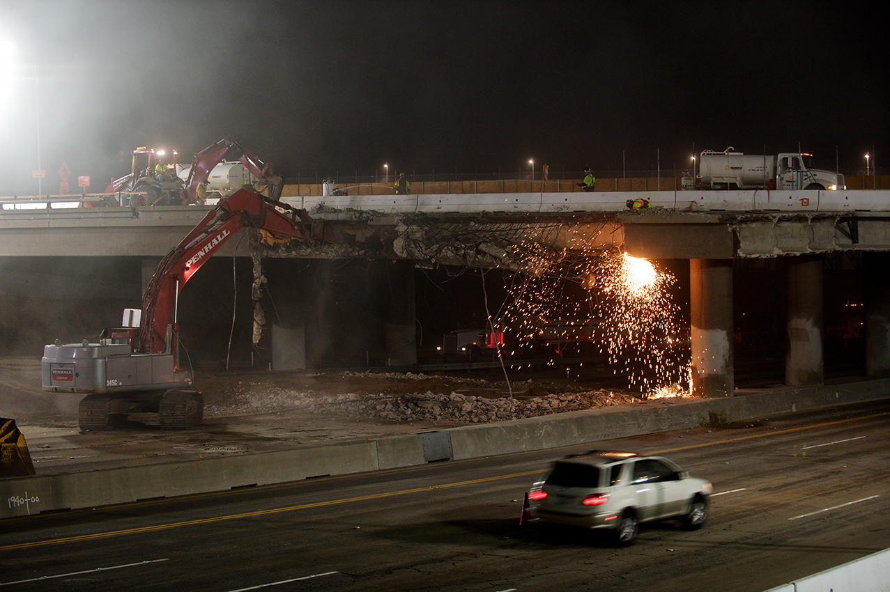 While a vehicle speeds south, the hoe ram punches at the north façade. Water trucks on the bridge spray water on the work area to reduce dust. Concrete from the bridge will be crushed on the project crusher sites and reused in the project. Photo by Peter Watkinson. While a vehicle speeds south, the hoe ram punches at the north façade. Water trucks on the bridge spray water on the work area to reduce dust. Concrete from the bridge will be crushed on the project crusher sites and reused in the project. Photo by Peter Watkinson.