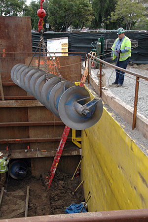 A worker guides a 24-foot-long auger as a crane lifts it from a work area. A worker guides a 24-foot-long auger as a crane lifts it from a work area north of Wilshire Bl. Another auger at the lower left waits to be removed. Chain rigging is used due to the sharp edges and irregular shape of the auger.