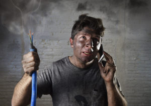 young man holding electrical cable smoking after electrical accident with dirty burnt face in funny desperate expression calling with mobile phone asking for help in electricity DIY repairs danger concept