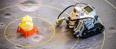 A Lego Mindstorms robot, with a plastic astronaut strapped to the front, approaches the lunar lander. Student teams will program the robot to explore the moon’s surface.