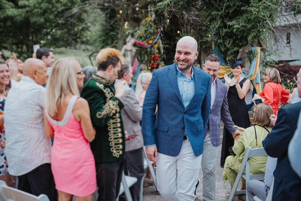 Two grooms hold hands as they leave the wedding altar, surrounded by cheering friends and family