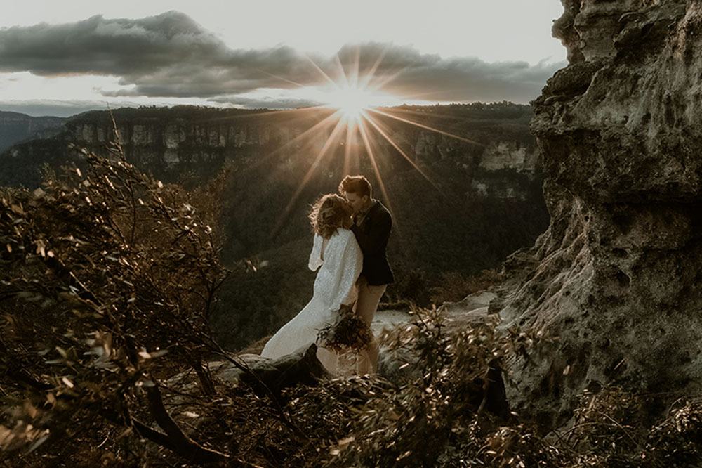Bride and groom embrace outdoors in front of mountains and a sunset