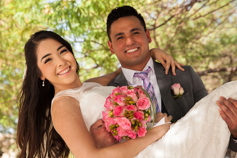 A groom and bride pose on the wedding day. He is holding her up in his arms, and she holds a bouquet of pink flowers. There is a tree behind them, and sunlight filters through the leaves for a sweet summer wedding photo
