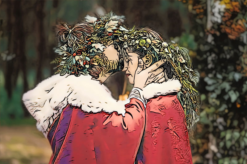 A groom and bride wearing red robes and wreaths of flowers on their heads pose on the wedding day. The groom cups the bride's face and kisses her on the forehead.