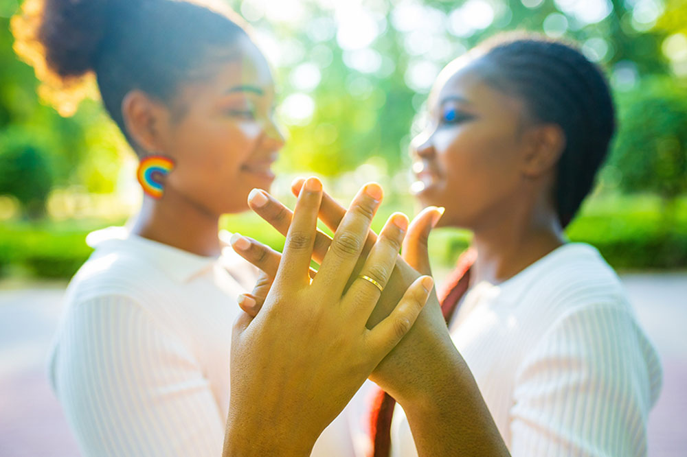 Close up photo shows two young brides holding out their hands to show off their wedding rings. One bride wears rainbow earrings.