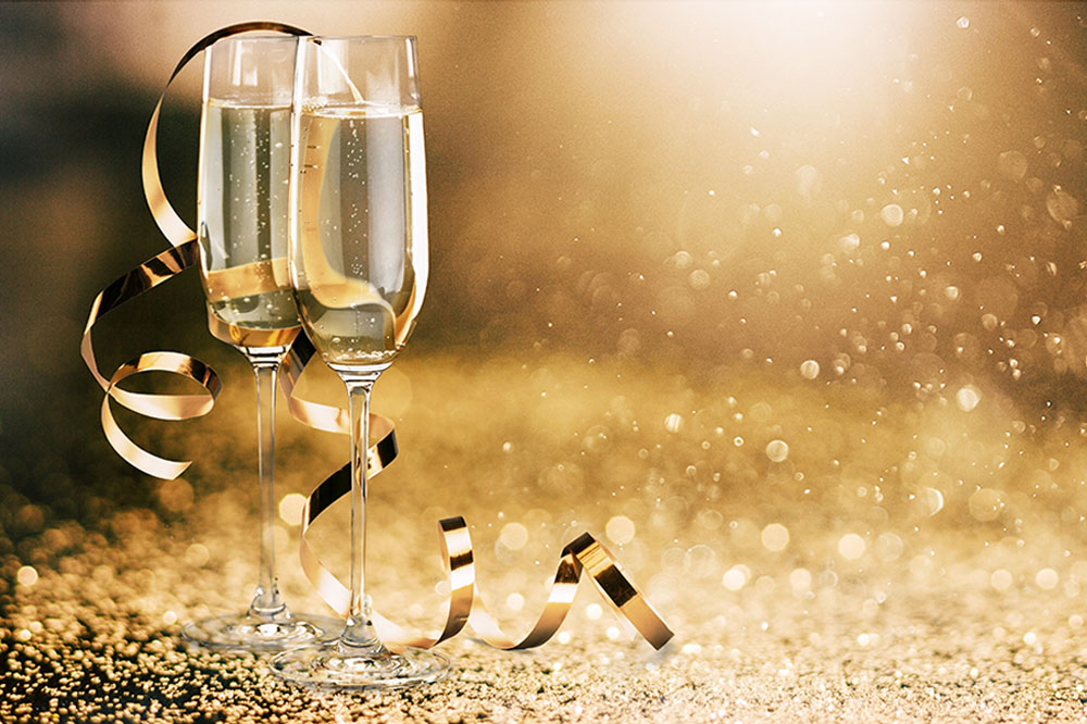 Two flutes of champagne sit on a table, with a strand of shiny golden ribbon curled and resting on the glasses. There is golden glitter on the table and falling all around in celebration.