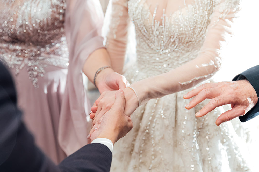 Close up photo shows two women in formal wedding clothing and two men in suits. They are all reaching forward to hold hands.