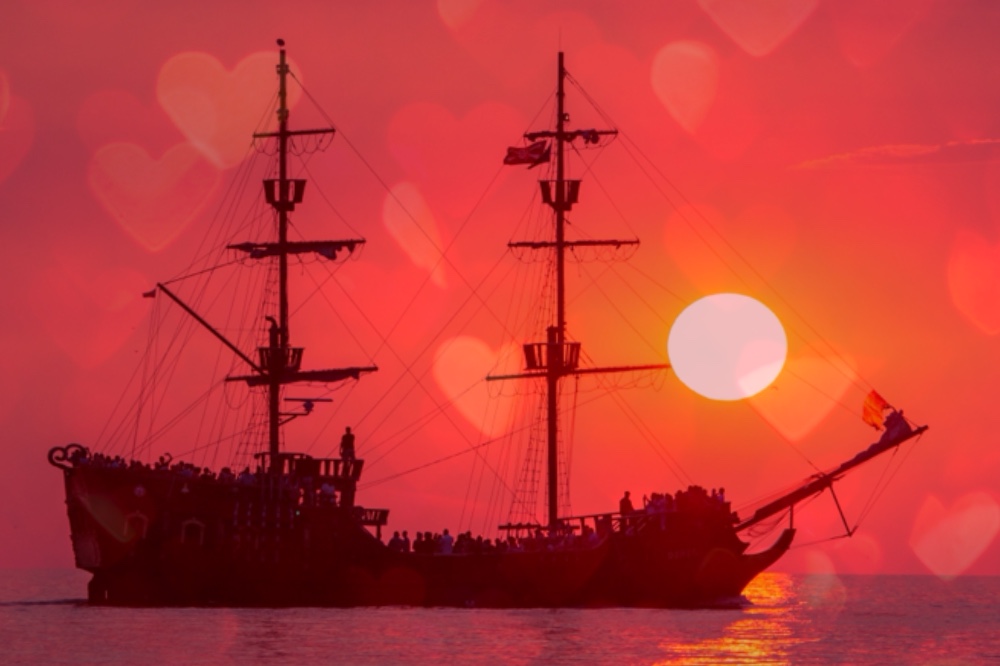 Photo shows a large pirate ship sailing at sunset, pink and red hearts have been super-imposed over the photo giving it a dreamy, romantic feel.