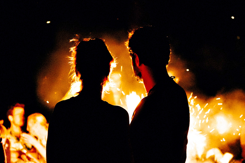 image is of a young couple, a man and woman, taken from behind as they stare into the glowing orange flames of a large beltane bonfire