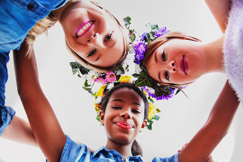 image is a photograph taken from below, looking up, at three smiling woman with floral wreaths on their heads in celebration of beltane