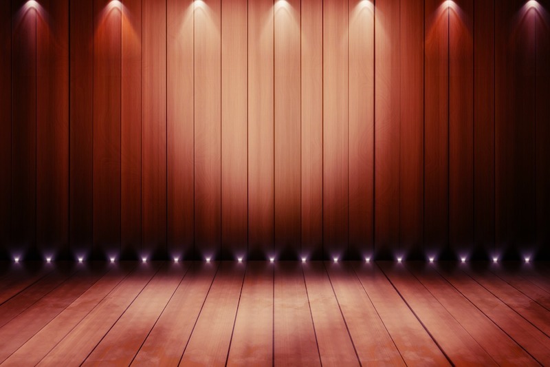 image is of an empty theater stage, representing the unknown of the wedding ceremony, there is a red fabric backdrop and lights shine on the wooden slats of the stage flooring 