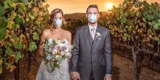 image of a bride and groom wearing masks outdoors in wedding clothing, while a wildfire fills the sky with orange and yellow smoke behind them