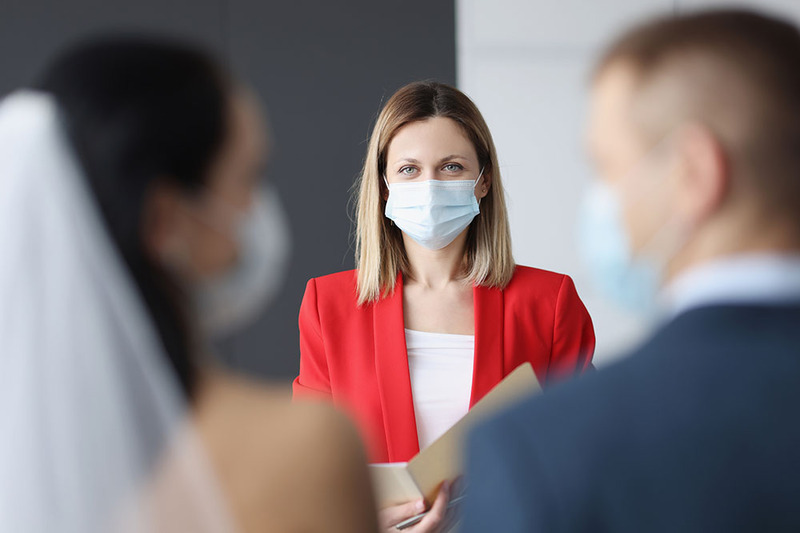 A wedding officiant wears a mask while officiating a ceremony, the photo is taken from behind the couple. The officiant wears a red jacket and her eyes show she is smiling behind the mask. 