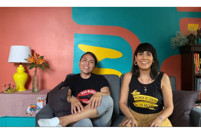 A photo of Karla and Daniela of Once Upon a Vow taken from their "about us" video. Karla is on the left, with her hair pulled back, wearing a black shirt with lettering that is partially obscured, that begins, "pro black" and smiling. Daniela is wearing a black tank top, with the lettering "women of color are the backbone..." and yellow pants. She is smiling. The room is painted and decorated in vibrant bright colors.