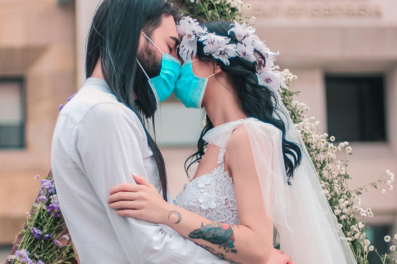 A young couple embrace during their wedding ceremony while wearing blue surgical masks. They both have long dark hair and are wearing white, and their foreheads rest together.