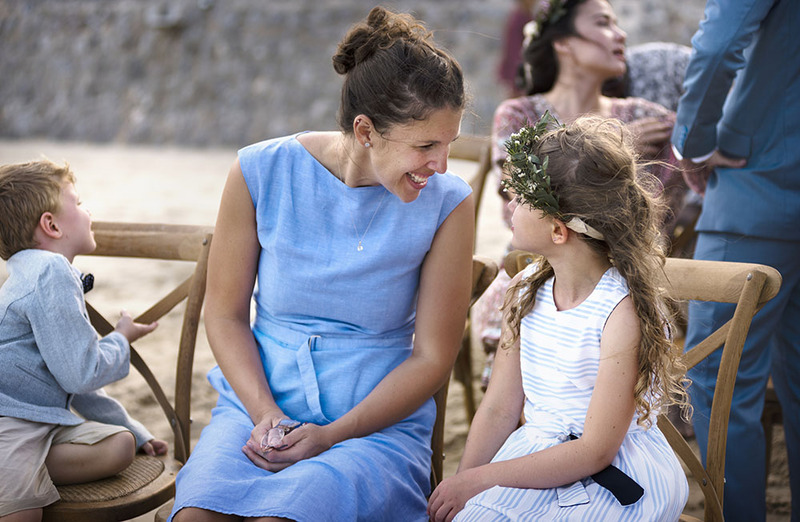 A wedding officiant sits next to a young flower girl at an outdoor wedding, chatting with her. The officiant is smiling and wears a light blue dress with cap sleeves, and holds her reading glasses in her lap. The flower girl is around 7 or 8 years old, and is wearing wreath with flowers over her long hair. 