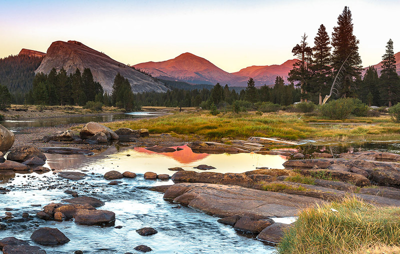 Yosemite park scene, mountains, pine trees, a river, and a beautiful sky behind at sunrise