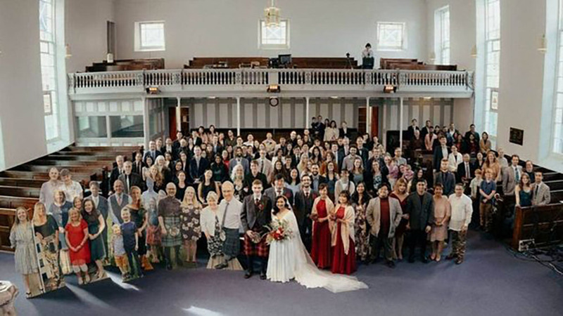 Photo via Daily Record, of the entire MacIver wedding guest list, including 20 cutouts, stand posing inside the wedding venue in New Zealand