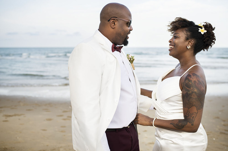  Un hombre y una mujer afroamericanos parados el uno junto al otro en la playa en el día de su boda, vestidos de  blanco. Están felices y ríen juntos.