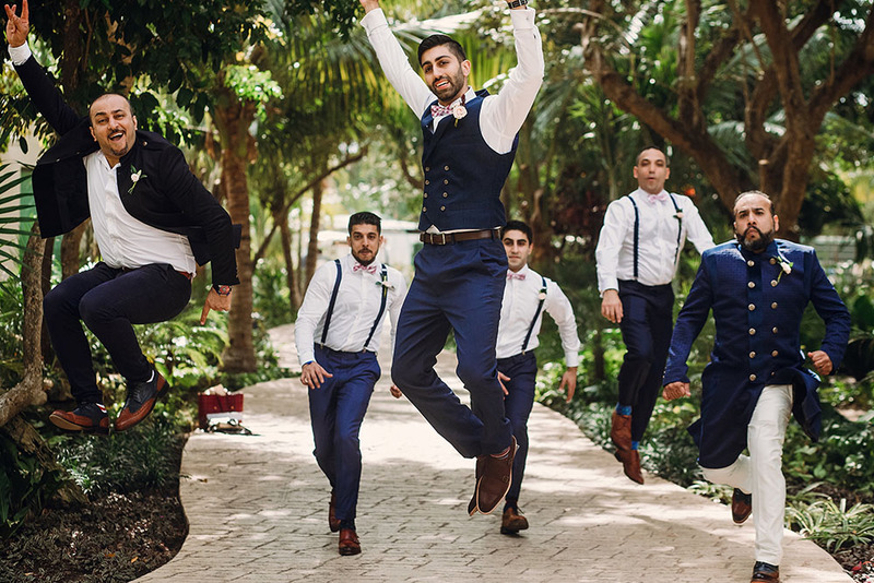 A group of groomsmen and a groom celebrate outdoors, some jump in the air