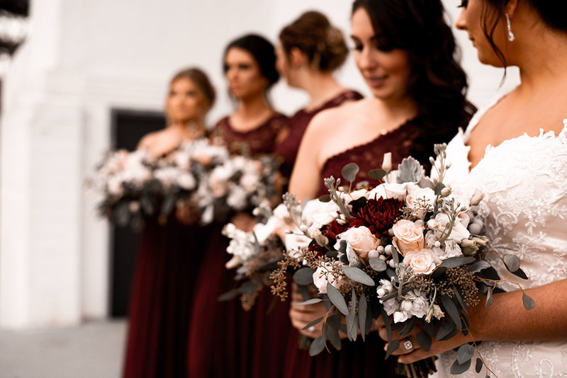 Bridesmaids lined up in a row with the bride in the front nearest the camera, they're holding flowers and wearing dark red dresses