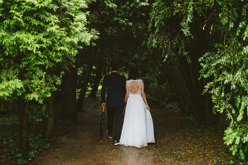 A groom and bride dressed in wedding clothes walk down a wooded path carrying an umbrella during an Oregon wedding ceremony