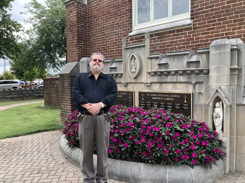 Image of Rev. Timothy Hooker, standing outside of a pretty brick building on a sunny day, wearing a black button up shirt with his hands folded in front of him, there are purple flowers and trees nearby. 