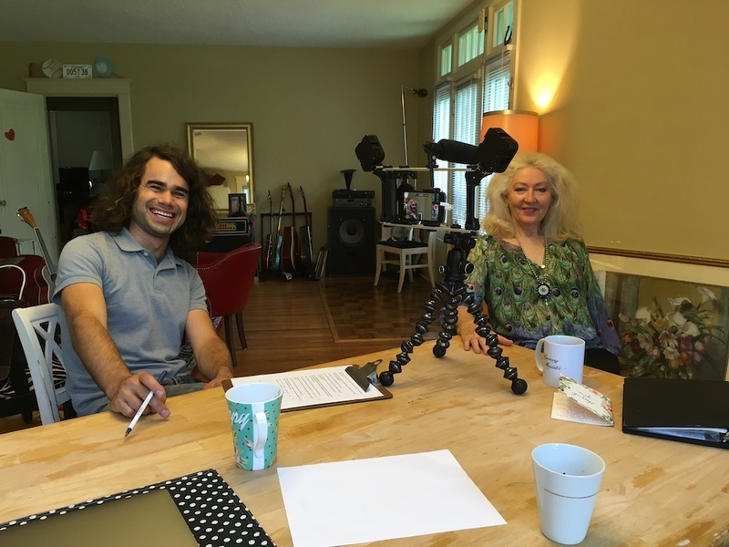 Image of AMM's Lewis King with AMM Minister Zelda Sheldon, they are sitting at a table smiling, Lewis is leaning back in his chair with a smile, his hair is long. Zelda sits beside him, wearing a patterned shirt and blonde hair to her shoulders.
