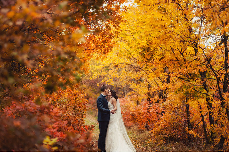 A bride and groom kiss outdoors in their wedding clothes surrounded by colorful orange and red fall tree leaves in Vermont