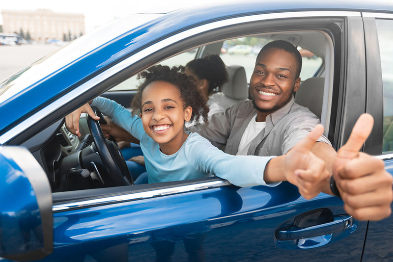 A happy family riding around in a car, the daughter and father face the camera and give the thumbs up sign