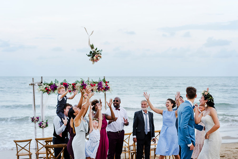 A happy group of friends and family celebrate a new marriage by tossing a wedding bouquet outdoors on the beach