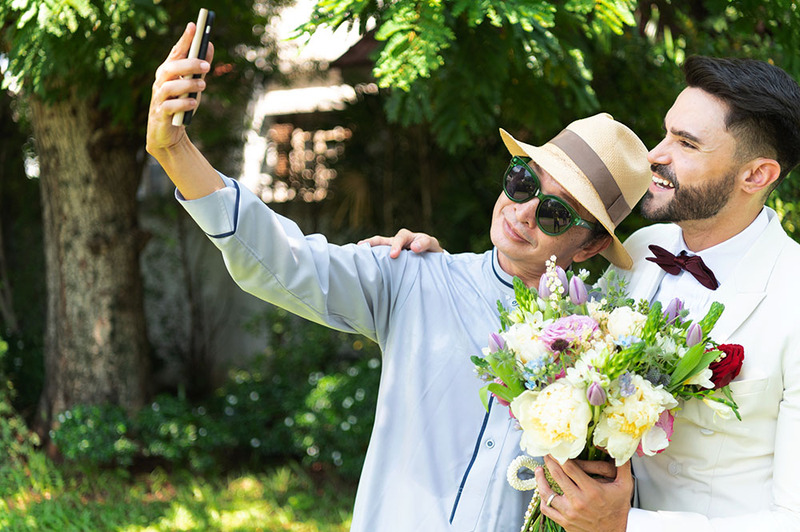 A young couple takes a selfie on their wedding day outdoors while holding flowers