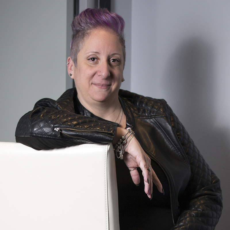 Wedding Officiant & Rabbi, Lori Prashker-Thomas headshot photo, shows Lori with short pink hair and a black leather jacket posing in a chair and smiling toward the camera