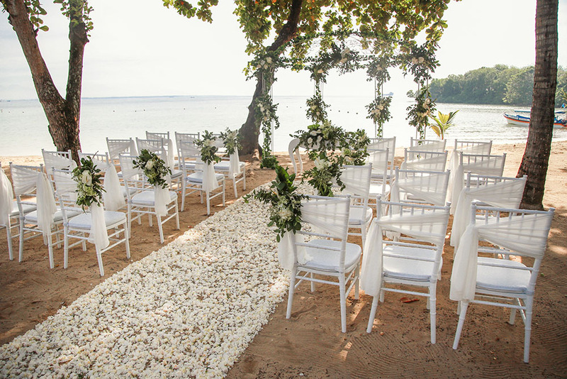 Chairs set up for a wedding ceremony on a beach, near the water, with white petals decorating the aisle
