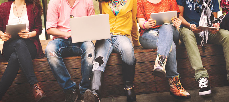 Photo shows a group of young people holding laptops and tablets, sitting outside in brightly colored clothing, sitting in a row