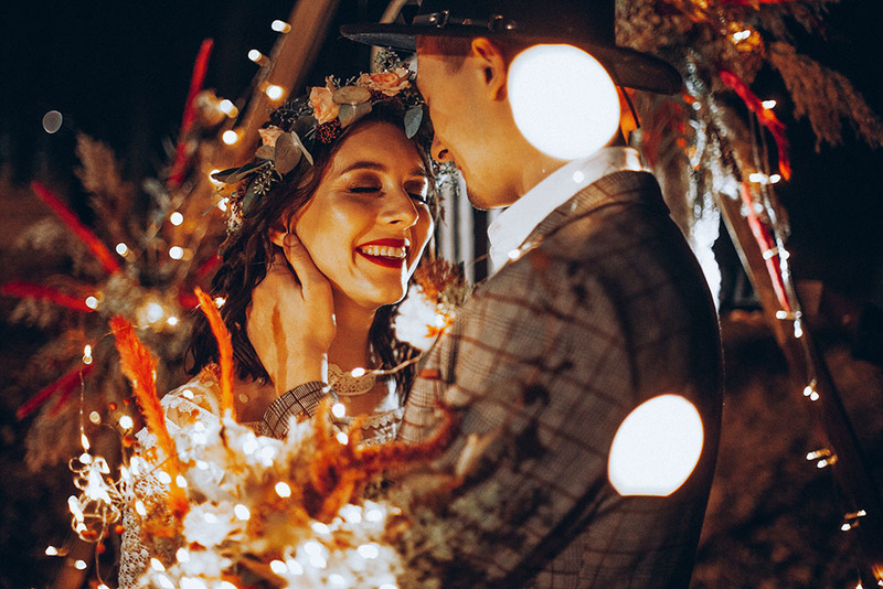 Happy newlyweds embrace following an outdoor ceremony at night, the arch behind them is lit up with soft twinkle lights, close up on their faces as they smile