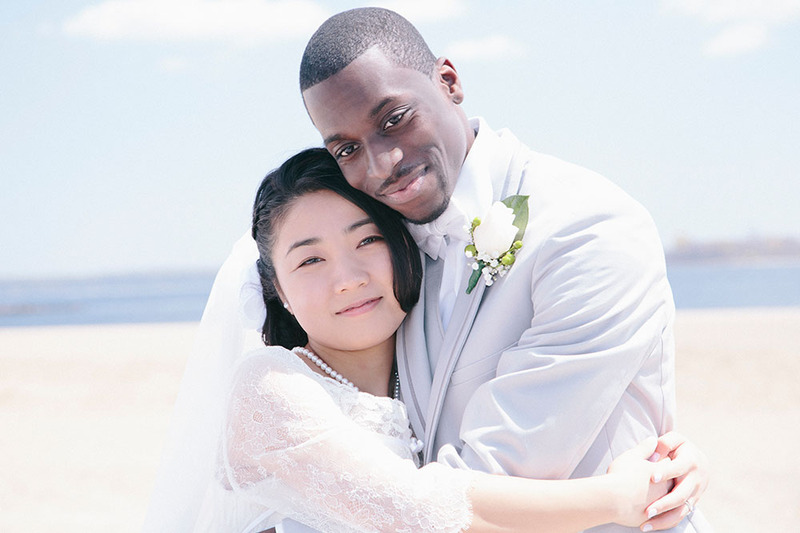 Newlyweds embrace, facing the camera, outside on the beach on a sunny day