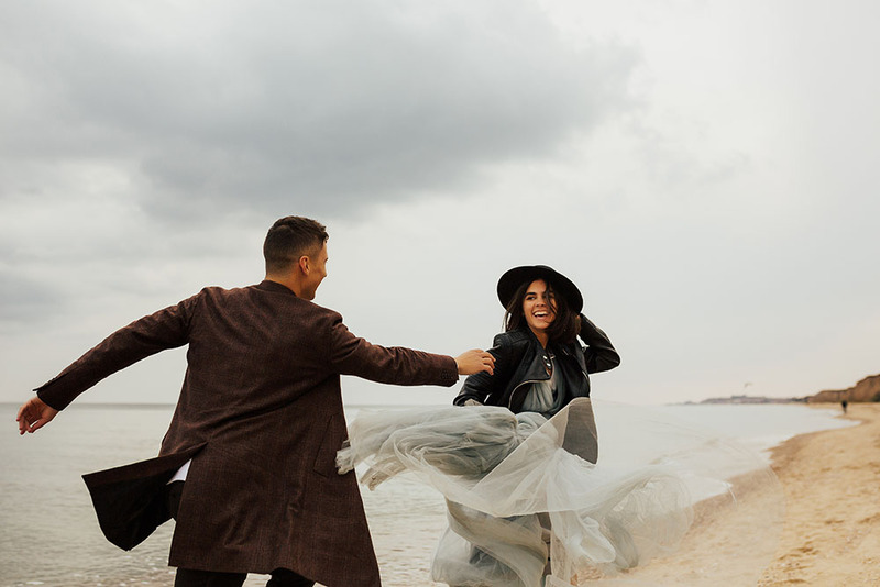 A young couple run down a beach in their wedding clothes, laughing and smiling, the bride turns to look back at the groom