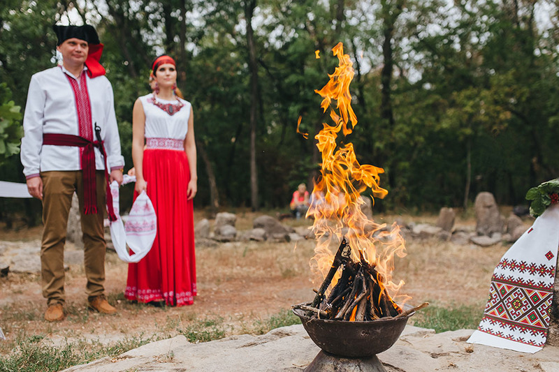 A Pagan couple celebrate their wedding outdoors, standing in front of a small bonfire with trees in the background, they are holding a handfasting cloth