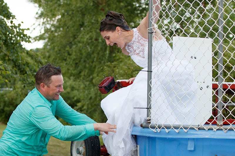 Screenshot from the YouTube video, Dunk Tank Wedding, showing the bride climbing out of the tank in her wet wedding dress while the groom watches, both are smiling and laughing happily