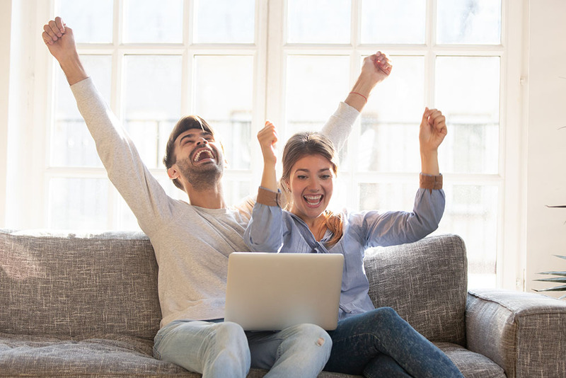 A happy couple sitting on a couch raise their arms and laugh in celebration of their new marriage, a laptop sits in their laps to represent the remote wedding officiant. They are dressed in casual clothing