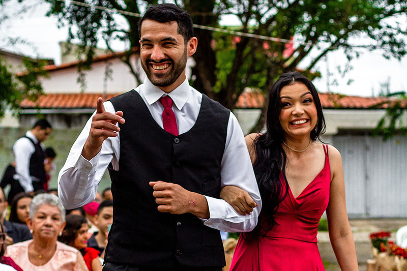 Photo of a happy bride and groom walking down the aisle outdoors at their wedding ceremony, the groom is in a suit and the bride is making a funny smile at the camera, she is wearing a red dress