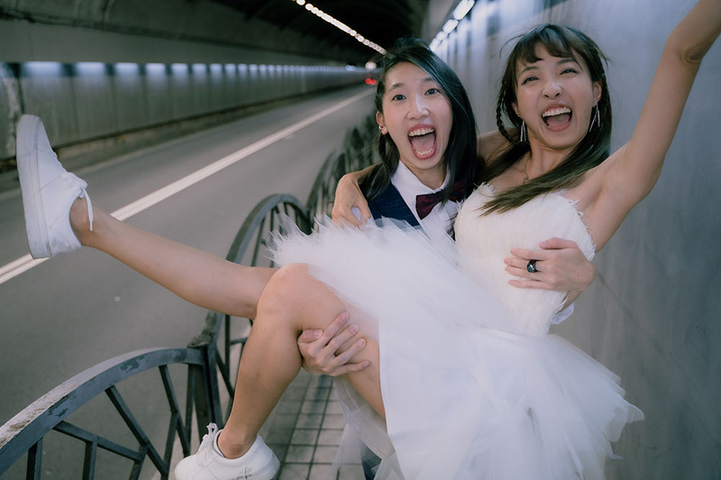 Two young brides celebrating their marriage, looking excitedly at the camera. One bride is wearing a suit, and holding the other, who is wearing a white dress, in her arms. 