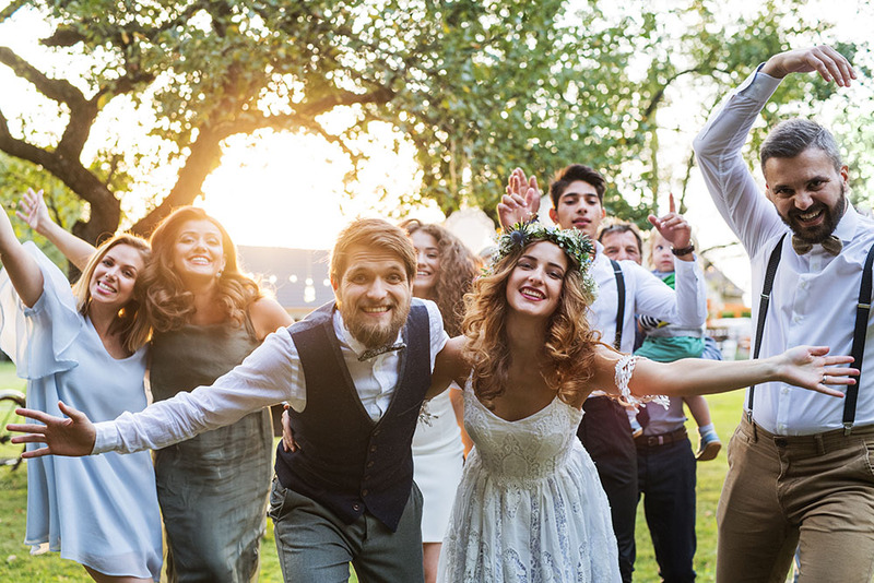 Two newlyweds hold out their arms and smile toward the camera after a sunny outdoor micro wedding ceremony, surrounded by friends