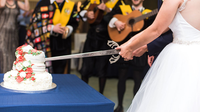 Close up photo of a bride and groom cutting a cake with a sword while friends and family watch in the background