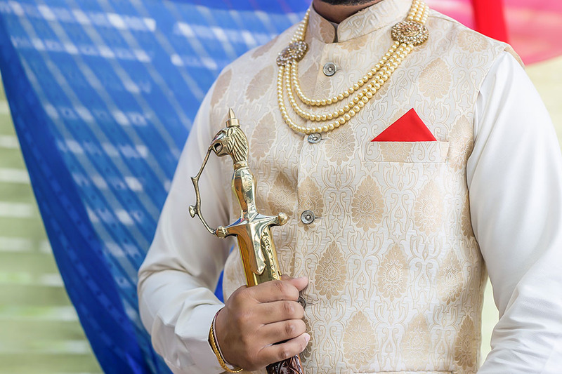 Close up photo of a man in a light colored suit with a red pocket square, holding a gold wedding kirpan