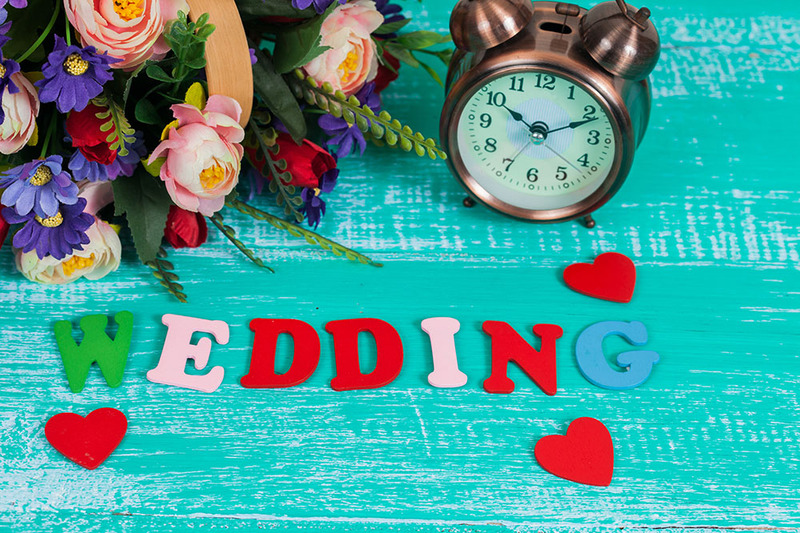 Stylized arranged photo of a flower bouquet and clock set on a teal background, with the word 'wedding' written in colorful wood block letters